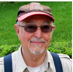 A man with a white beard, a baseball hat, and a pair of sunglasses smiles at the camera